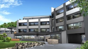 Un edificio moderno de varios pisos con techo plano, ventanas grandes y un estacionamiento con muro de piedra y vegetación.