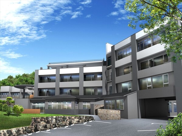 Un bâtiment moderne à plusieurs étages, doté d’un toit plat, de grandes fenêtres et d’un espace de stationnement agrémenté d’un mur en pierre et de verdure.