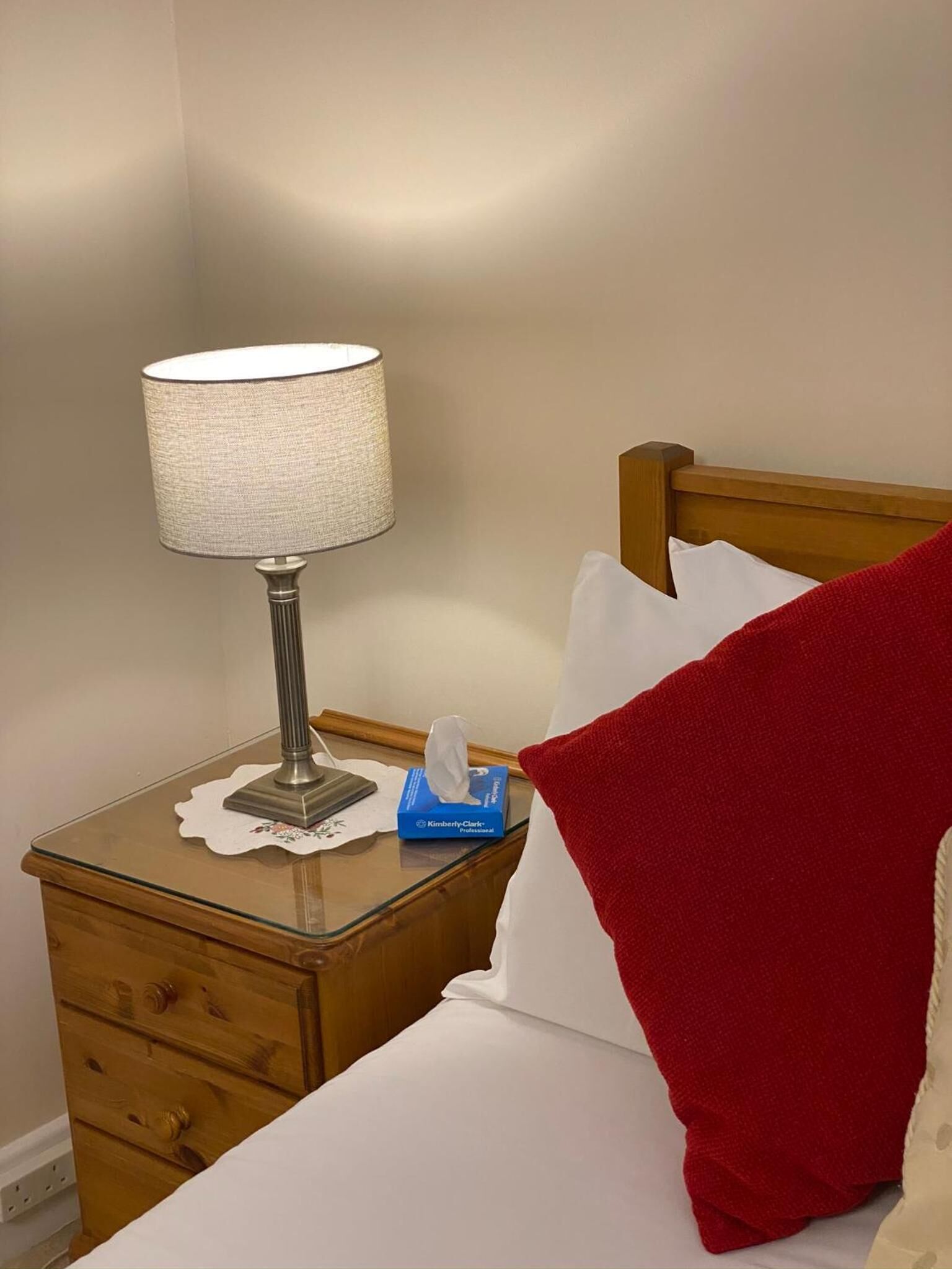 Una cama bien hecha con un almohadón rojo, una mesita de noche con una lámpara y una caja de pañuelos.