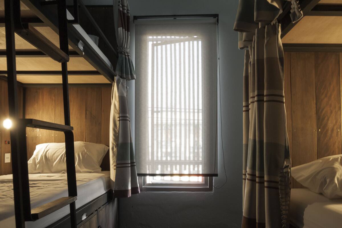 A dormitory room with bunk beds, a window with blinds, and a wooden floor.