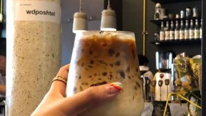 A hand holding a glass of iced coffee with a label on a pillar. - Whereder Poshtel - Hostel (Udon Thani)
