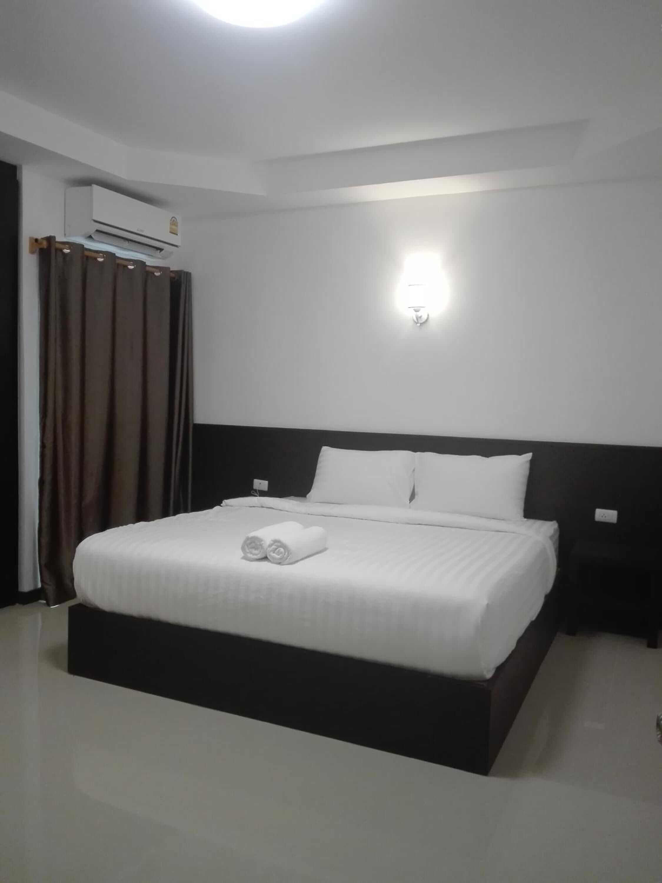 A neatly made bed with white linens and rolled towels, a dark headboard, and a wall-mounted light fixture.