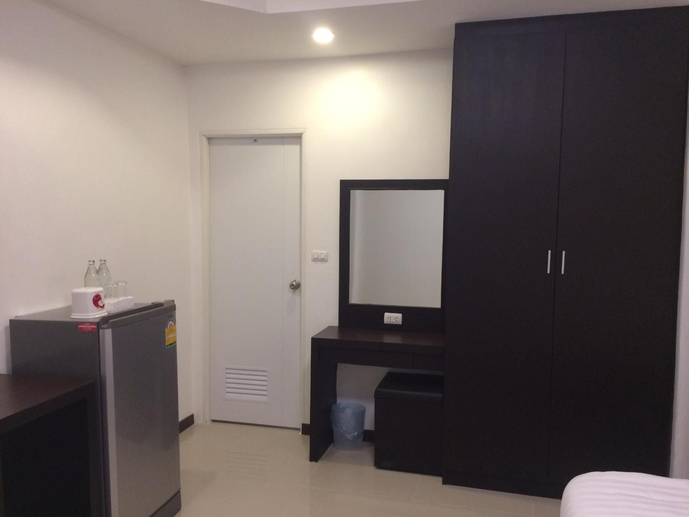 A small, modern hotel room with a bed, a desk with a mirror, a wardrobe, a refrigerator, and a trash bin.