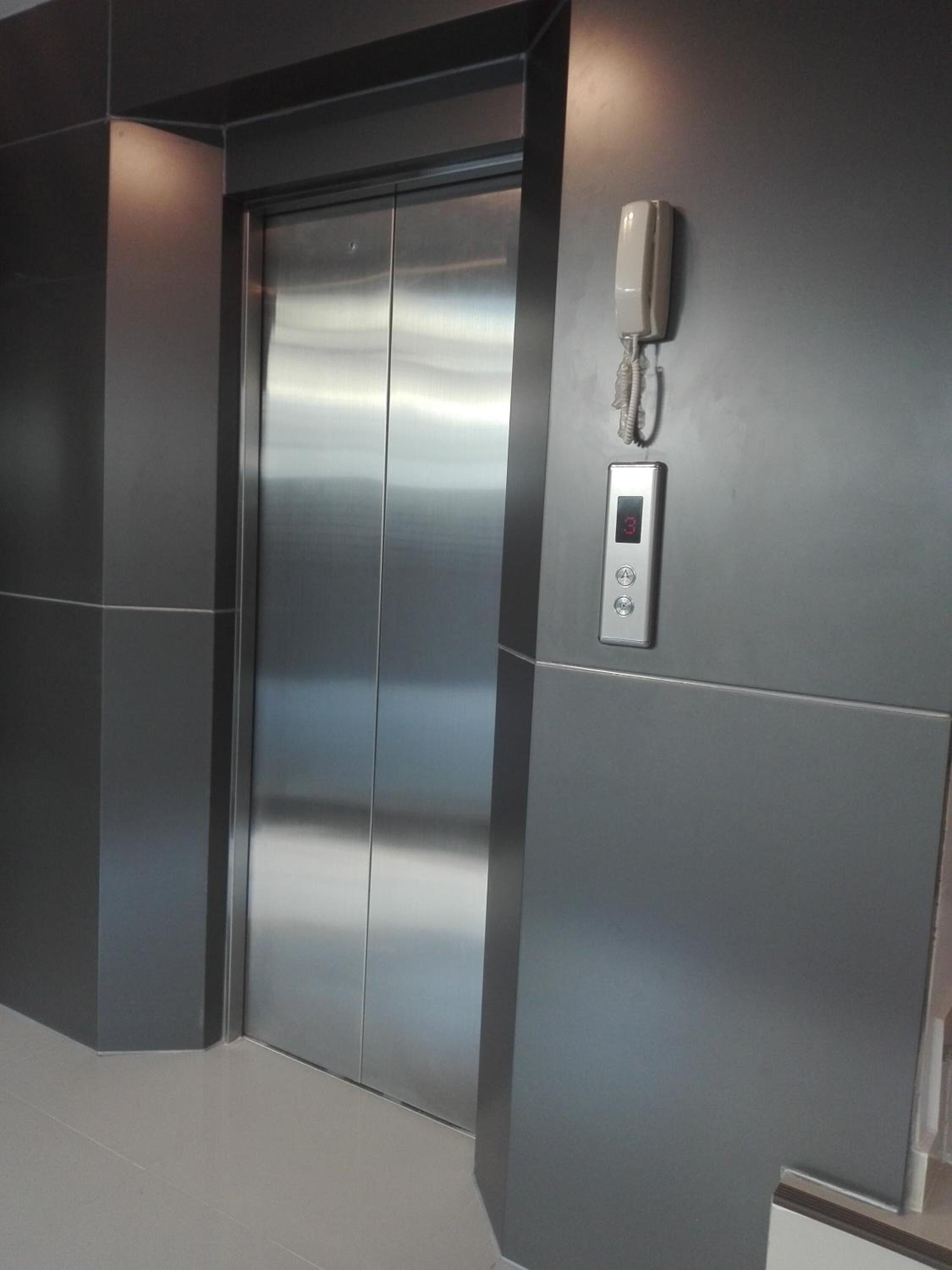 Een moderne lift met een digitaal scherm, een telefoon en een bedieningspaneel aan de muur.