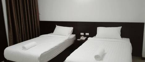 A hotel room with two single beds, each with a white bedspread and a rolled towel. There is a nightstand with a phone and a lamp between the beds.