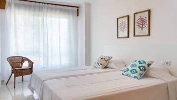A hotel room with two beds, a wicker chair, and two framed botanical prints on the wall.