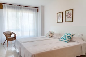 A hotel room with two beds, a wicker chair, and two framed botanical prints on the wall.