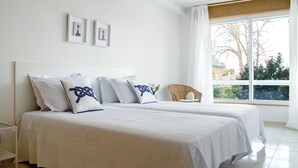 Un dormitorio con dos camas, ropa de cama blanca y un ventanal con vistas a árboles y un edificio.