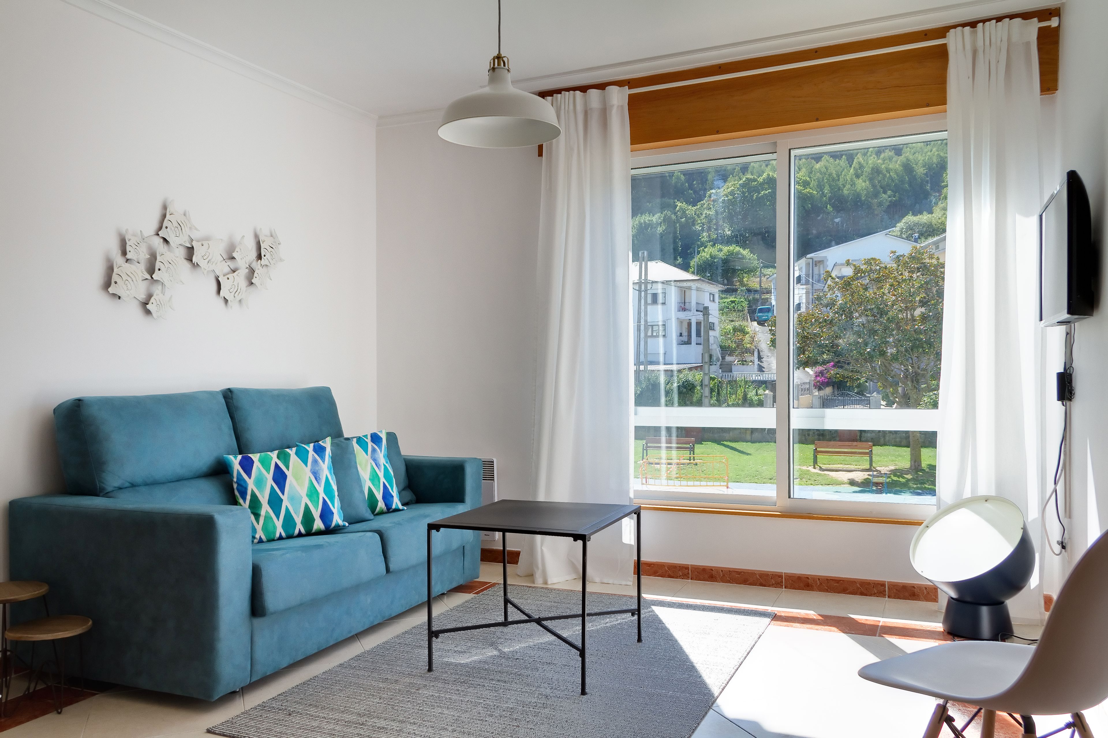Een moderne woonkamer met een blauwe bank, een kleine tafel en een stoel. Er is een groot raam met uitzicht op huizen en bomen.