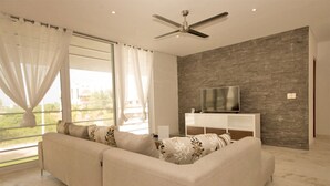 Ein modernes Wohnzimmer mit einem beigefarbenen Sofa, einem Flachbildfernseher auf einer weißen Konsole, einem Deckenventilator und einem großen Fenster mit durchsichtigen Vorhängen.