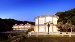 A large, circular building with a white exterior and multiple windows, surrounded by a white fence and palm trees.