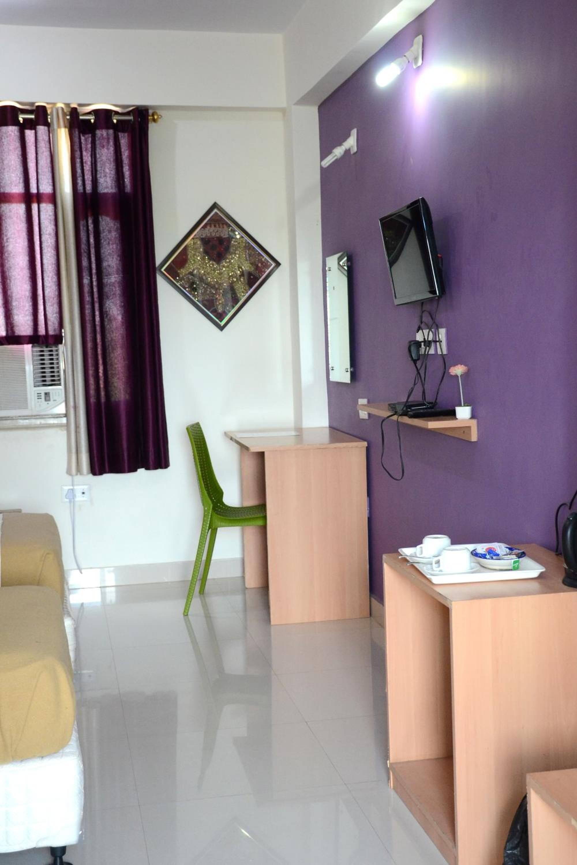 A room with a purple wall, a flat-screen TV mounted on it, a small wooden desk with a cup and saucer, and a green chair.