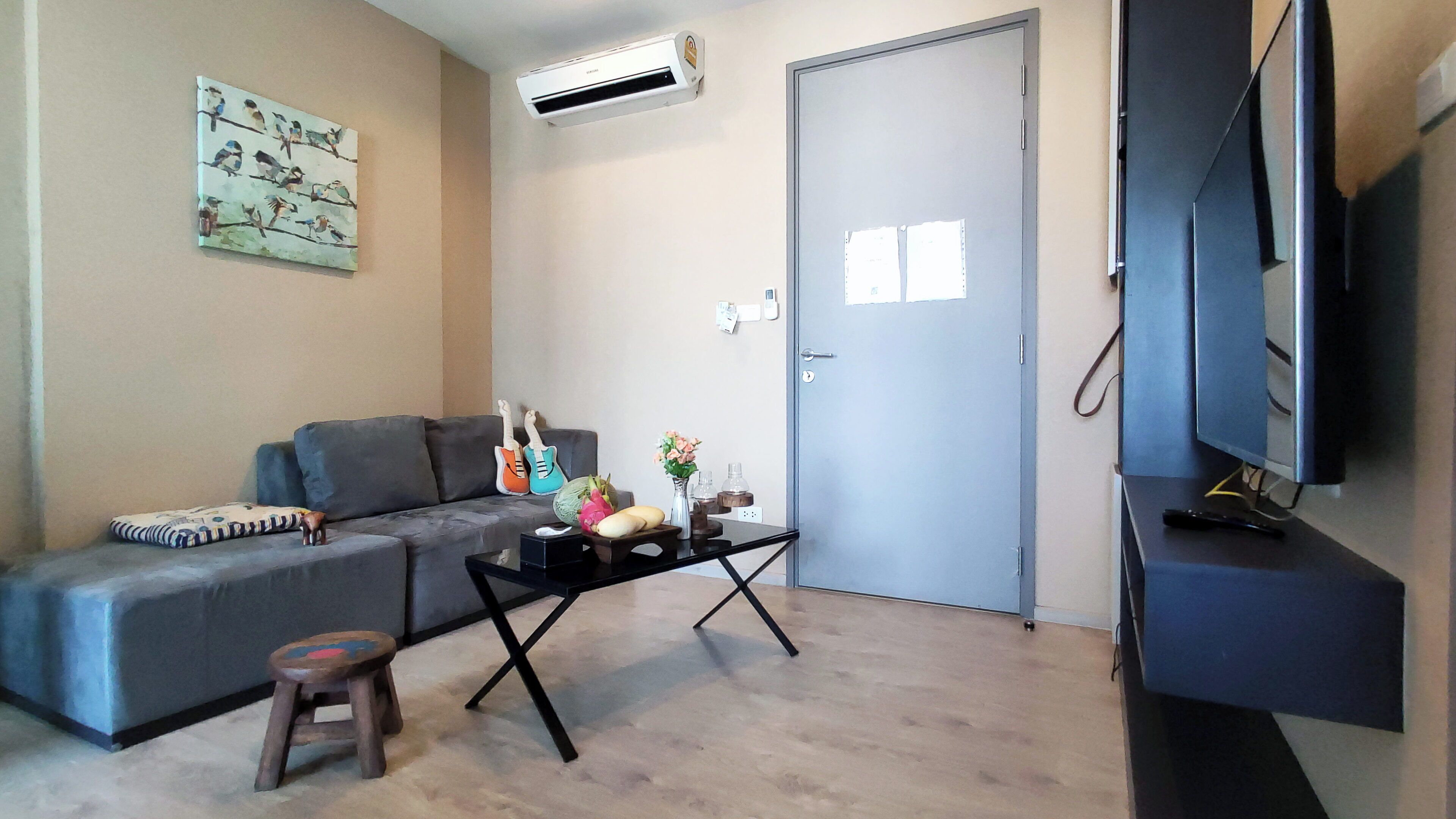 A living room with a grey sofa, a small wooden stool, a coffee table, and a flat-screen TV on a stand.