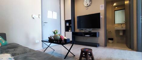 A modern living room with a flat-screen TV, a sofa, a small stool, and a dining table with a vase of flowers.