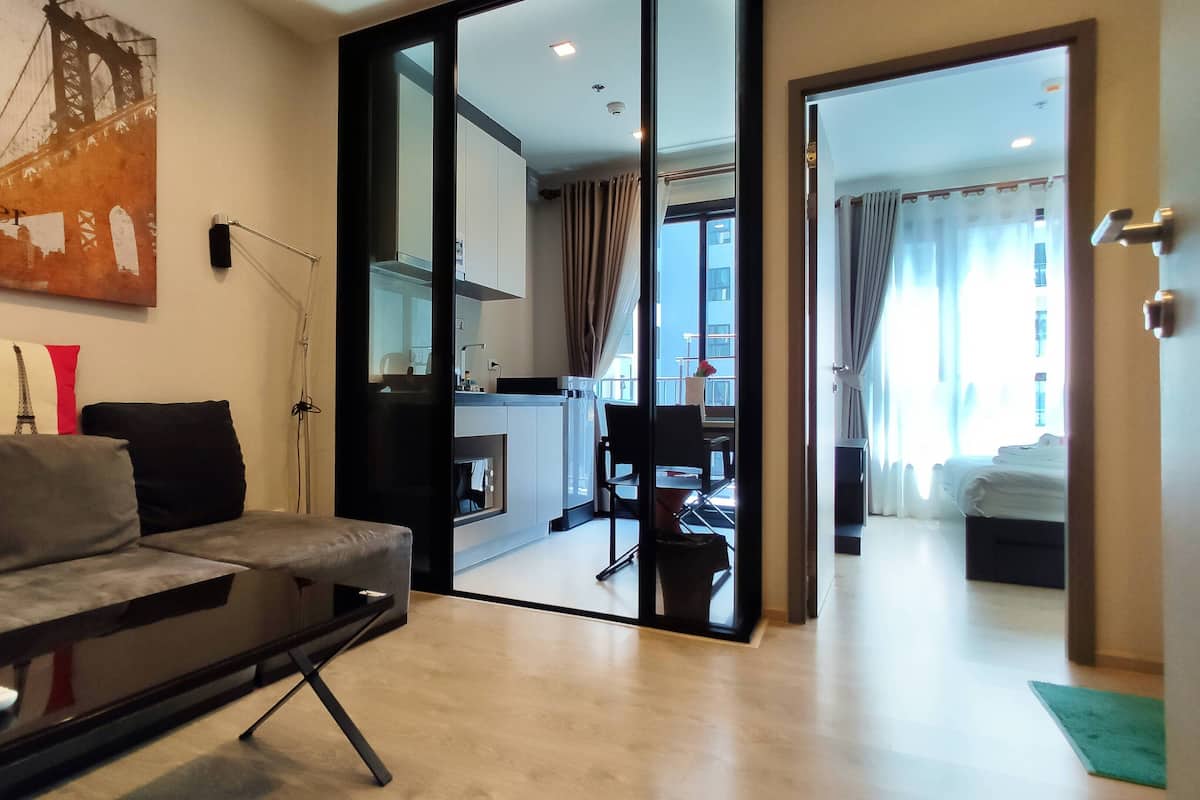 A modern living room with a sofa, a coffee table, and a painting of a bridge. There is a glass door leading to a kitchen and a dining area.