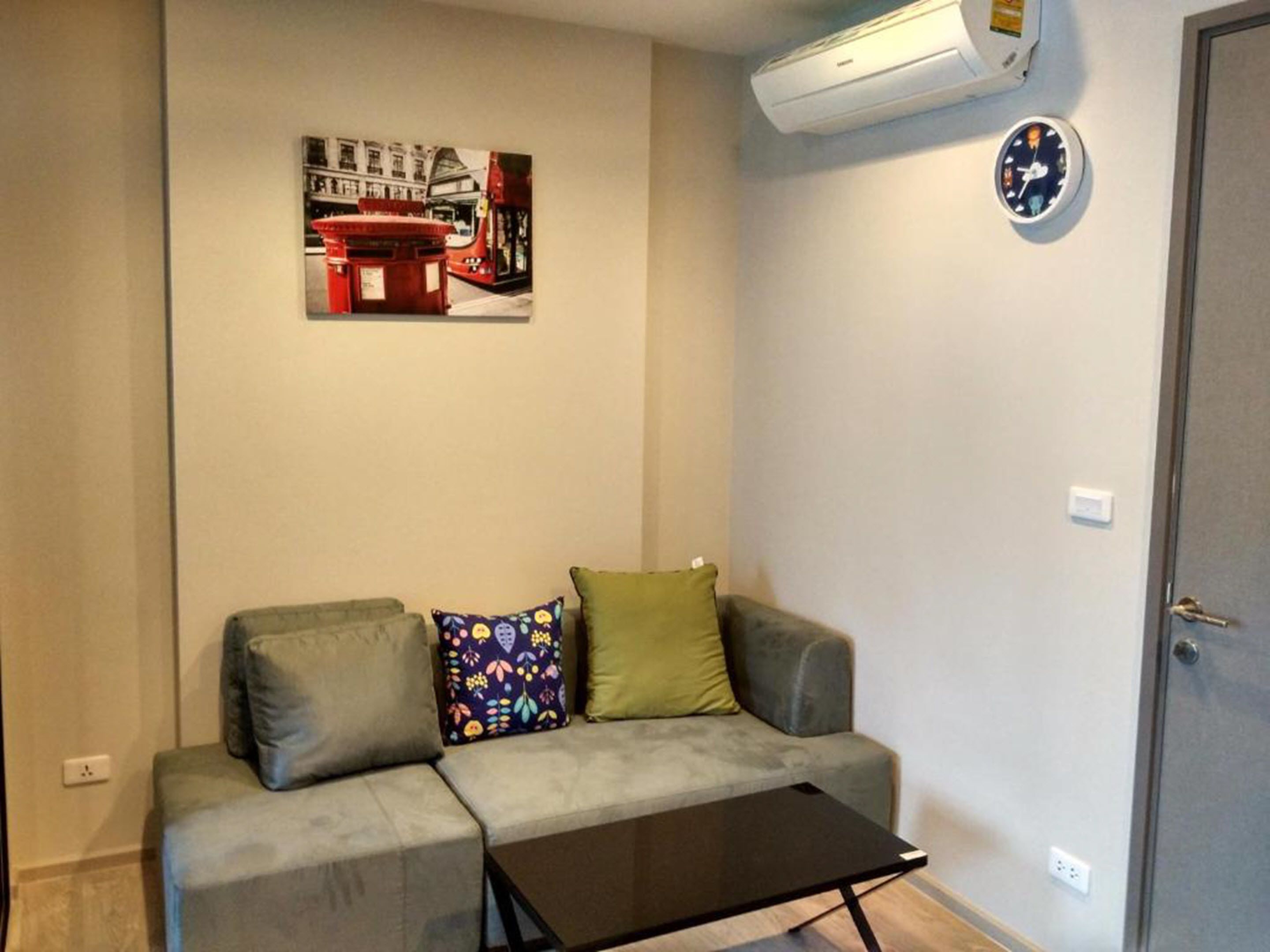 En stue med en grå sofa, en farverig pude, et sort sofabord, en fastmonteret aircondition og et ur.