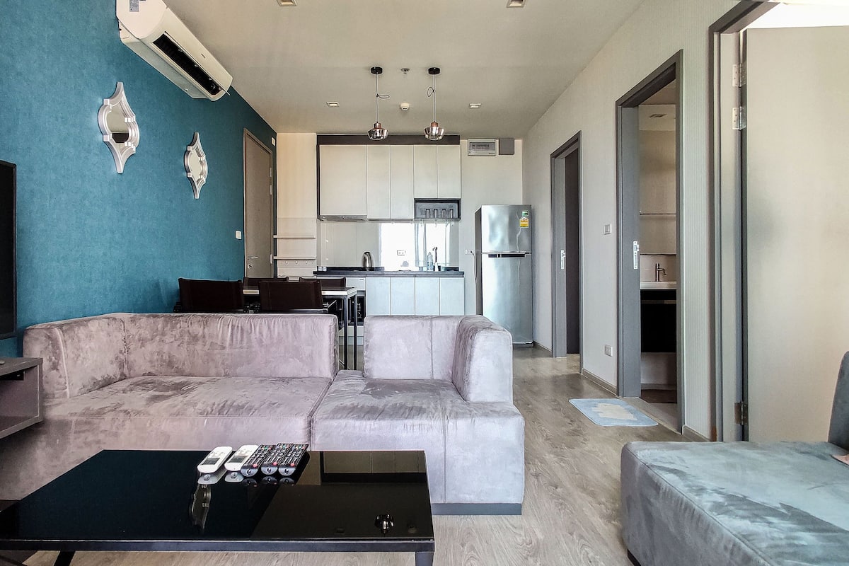 A modern living room with a sofa, a coffee table, and a TV. There is a kitchen in the background with a refrigerator and cabinets.