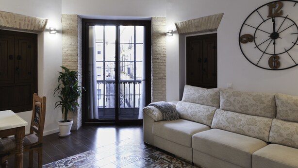 A living room with a patterned floor, a sofa, a dining table with chairs, a potted plant, and a large wall clock. - Posada Pilatos (Seville)