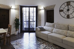 A living room with a patterned floor, a sofa, a dining table with chairs, a potted plant, and a large wall clock. - Posada Pilatos (Seville)