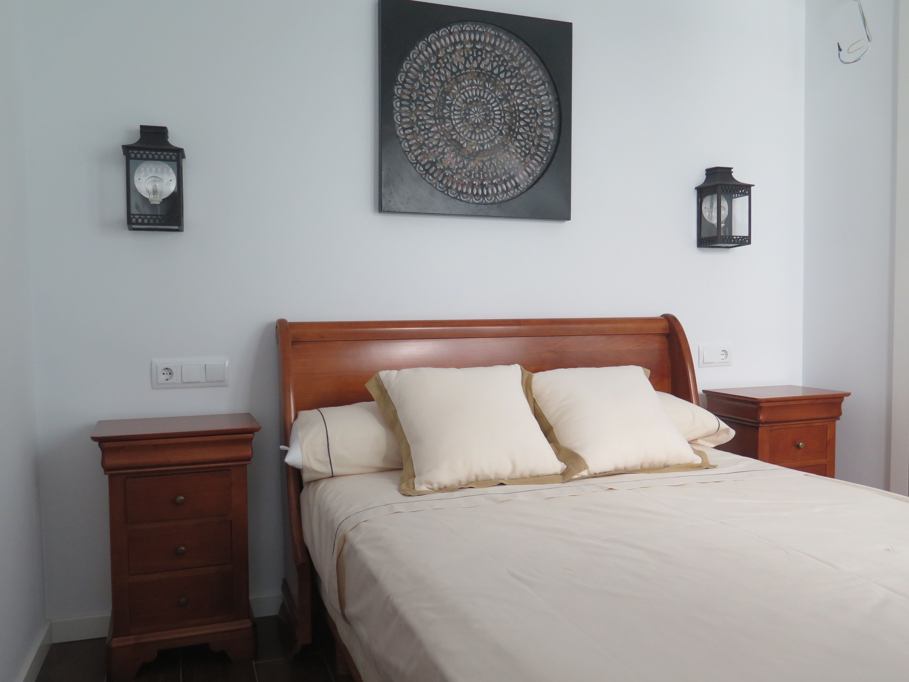 Un dormitorio con una cama de madera, dos mesitas de noche, un adorno en la pared y dos faroles de pared.