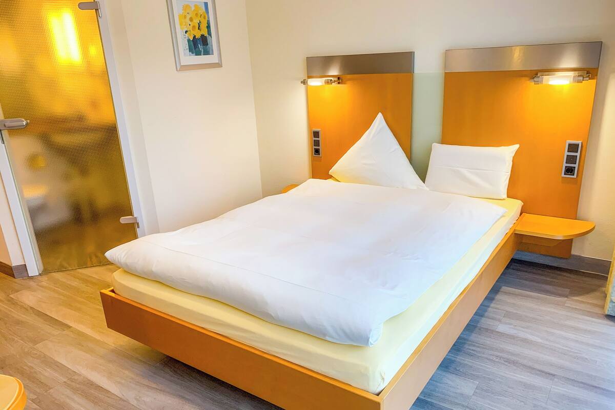 A hotel room with a bed, wooden headboard, bedside tables, and a wall-mounted light fixture.