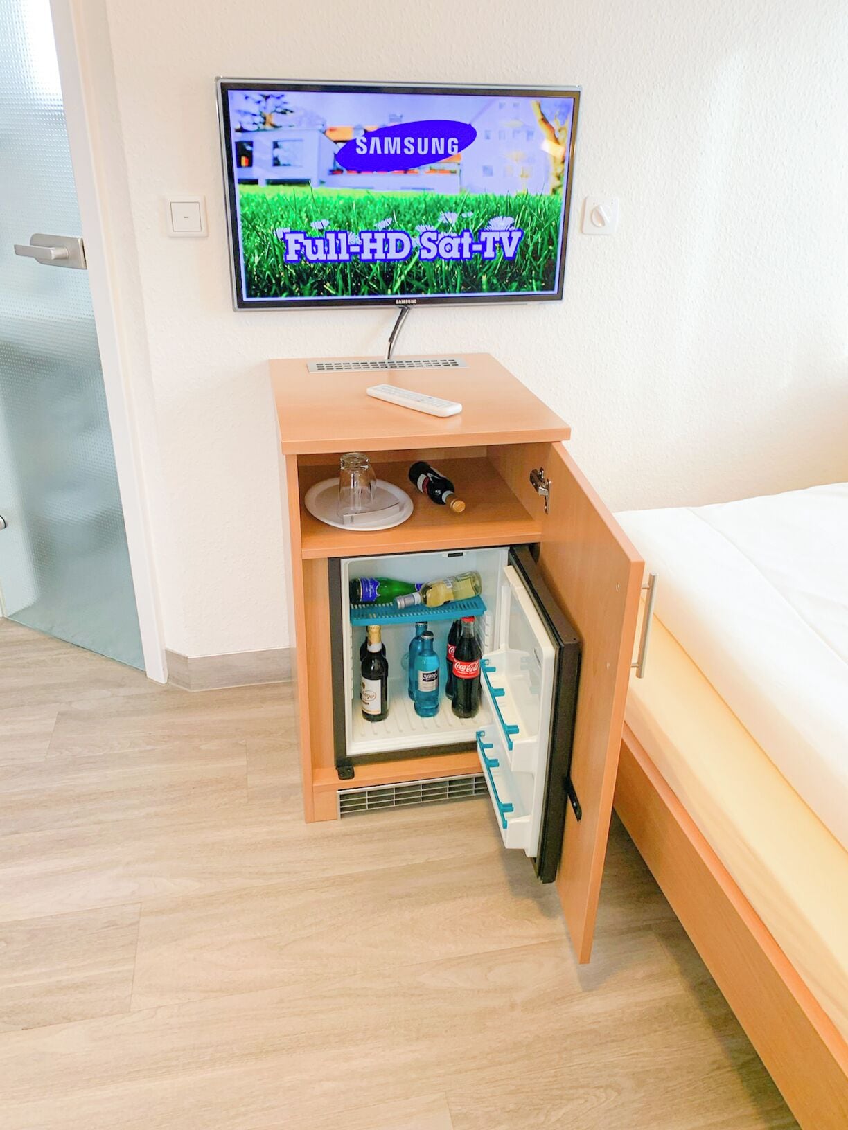 A room with a bed, a small wooden cabinet with a television mounted above it, and a mini-fridge with beverages.