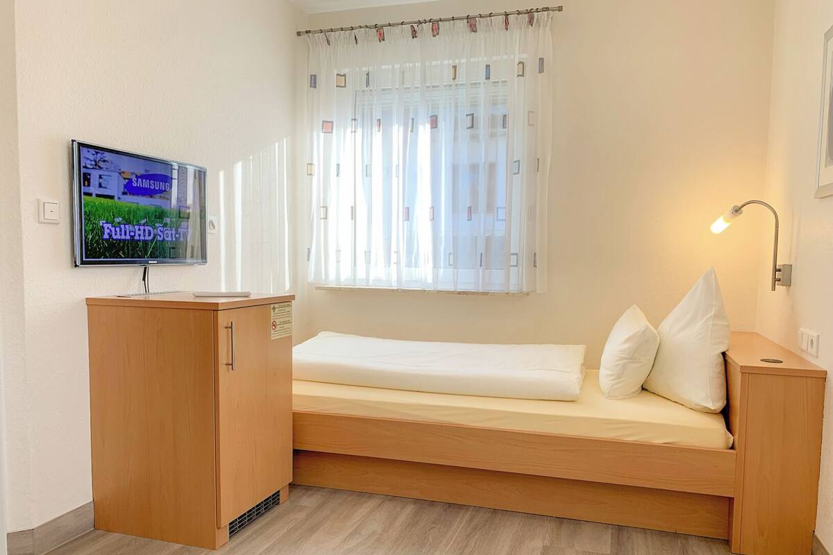 A small, single-bed room with a wooden desk, a television mounted on the wall, and a bedside lamp.
