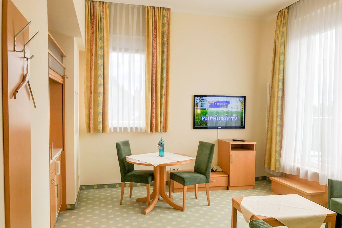 A hotel room with a dining area, a TV displaying 'Full-HD Sat-TV', and a window with curtains.