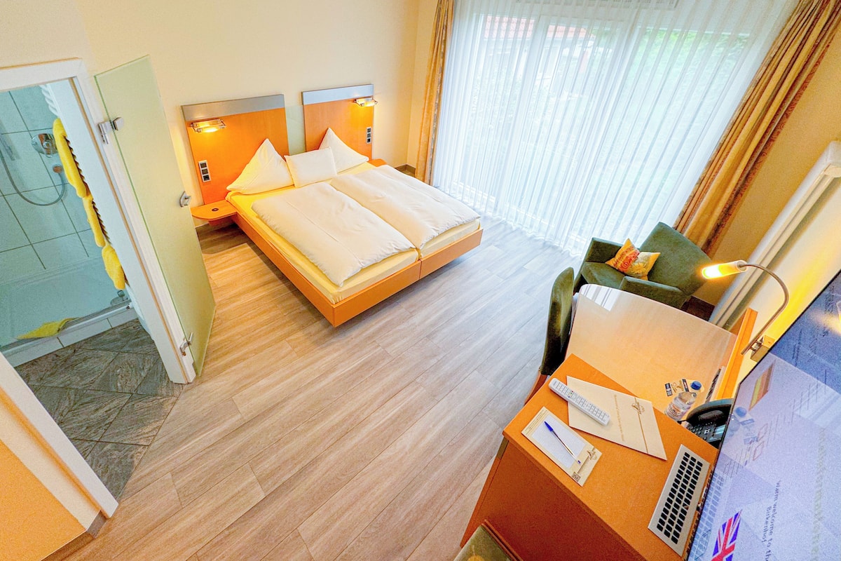 A hotel room with a double bed, a desk with a chair, a shower area, and a large window with curtains.