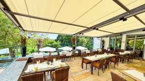 Een buitenterras met tafels en rieten stoelen, overdekt door een zonnescherm en omringd door glazen wanden.