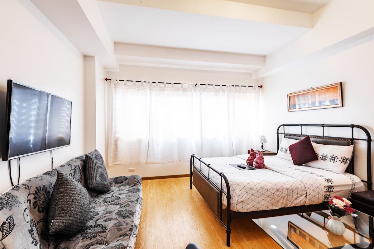 A bedroom with a bed, a sofa, a TV, and a window with curtains.