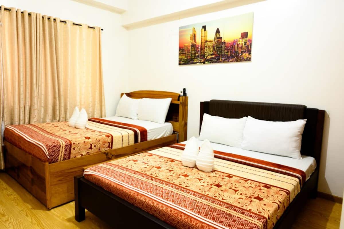 A hotel room with two beds, each with a wooden headboard, white bedding, and a patterned red and beige bedspread. A cityscape painting is on the wall.