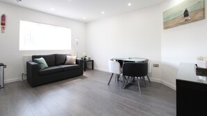 Una sala moderna con un sofá gris oscuro, una mesa de comedor de vidrio y una silla blanca. Hay un ventanal con persianas, una mesita auxiliar con una lámpara y un cuadro en la pared.
