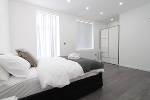 A modern bedroom with a bed, a radiator, a wardrobe, and a window with blinds. - BlueGreen Suites (Edgware)