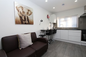 A modern living room with a brown sofa, a painting of an elephant, a kitchen with white cabinets, and a window with blinds.