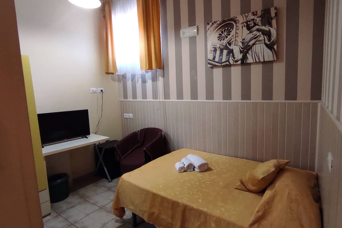 Pieni hotellihuone, jossa on yksi sänky, työpöytä tietokoneella, tuoli ja seinälle ripustettu taideteos.