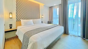 Quarto de hotel com uma cama grande, cabeceira estampada, uma mesa pequena e vista para o exterior através de amplas janelas.