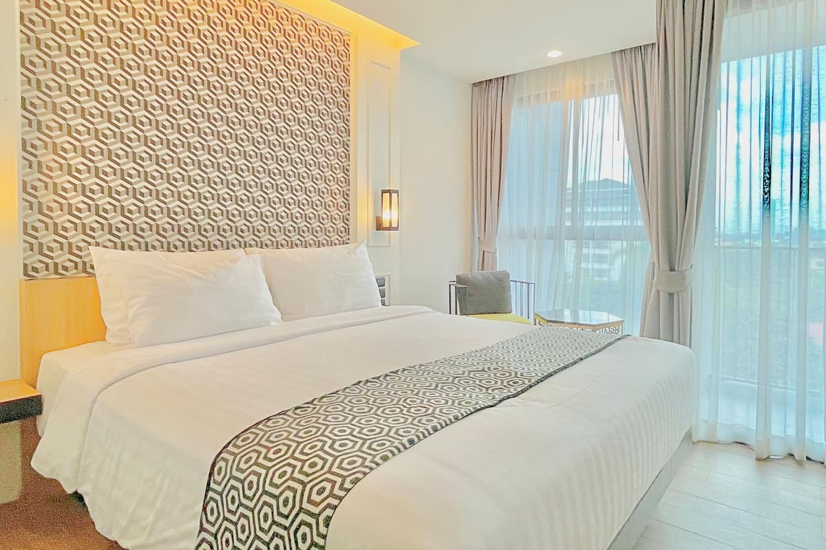A hotel room with a large bed, a geometric patterned headboard, a small table with a chair, and a view of the outdoors through sheer curtains.