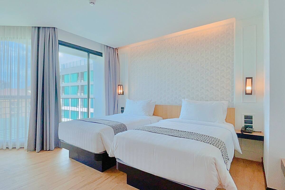 A hotel room with two beds, a large window with curtains, a patterned wall, and a nightstand with a phone.
