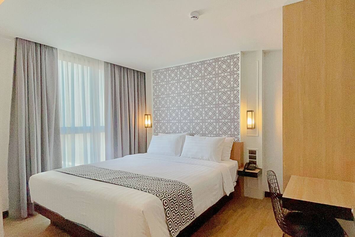 A hotel room with a large bed, a patterned wall, a nightstand, a chair, and a window with curtains.
