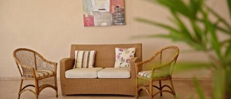 Un sofá de mimbre con cojines y dos sillones de mimbre en una habitación con suelo de baldosas y un cuadro abstracto colgado en la pared.