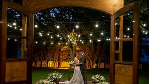 Un arco de madera que conduce a un espacio para una ceremonia de boda, con luces colgantes y arreglos florales.