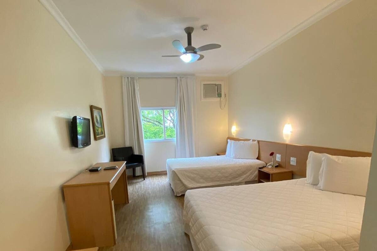 A hotel room with two beds, a desk, a chair, a ceiling fan, and a window with curtains.