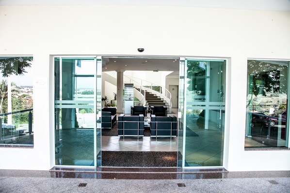 Una entrada de edificio moderna con puertas de vidrio, una escalera que conduce a un nivel superior y un área de estar con sillas negras y una mesa de centro.