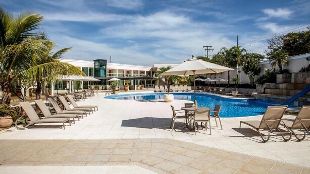 Zona de piscina con sillones de descanso y sombrillas, rodeada de palmeras y un edificio con un letrero verde.
