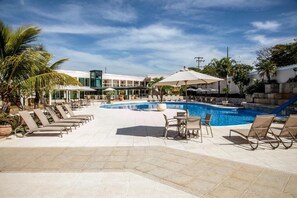 Zona de piscina con sillones de descanso y sombrillas, rodeada de palmeras y un edificio con un letrero verde.