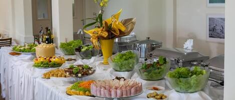 A buffet table with various food items, including salads, meats, and a cake, in a well-lit room with a white tablecloth.