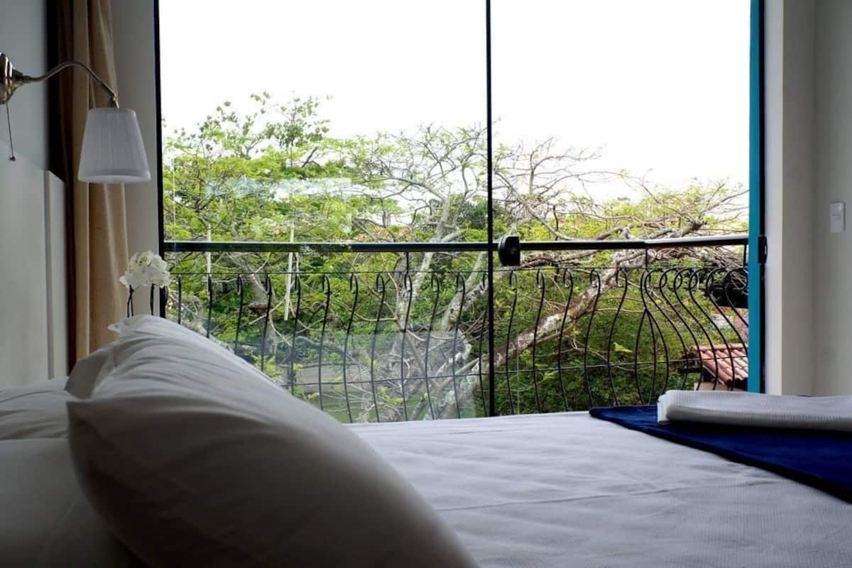 Ein Hotelzimmer mit großem Fenster, einem Bett mit weißer Bettwäsche, einem Metall-Balkongeländer und Blick auf Bäume sowie ein Gebäude.