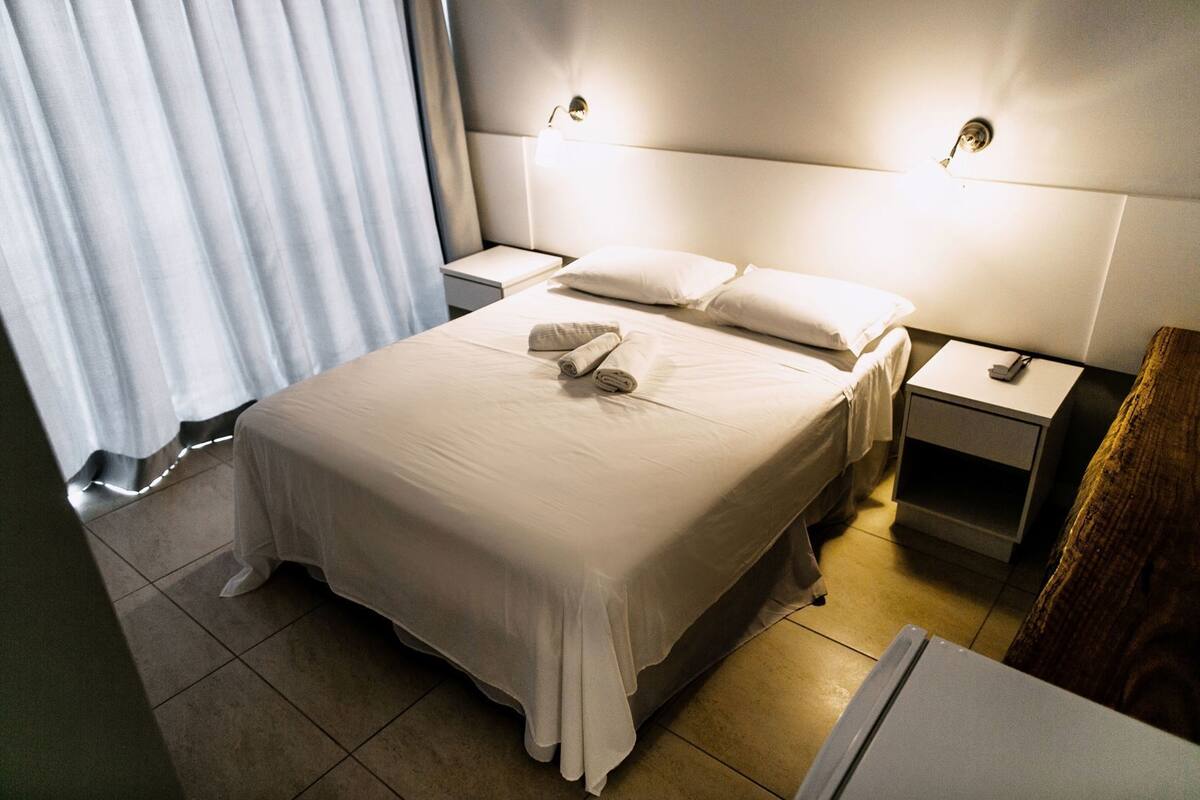 Ein Hotelzimmer mit einem großen Bett, zwei Nachttischen, einem Beistelltisch, einem Holzstuhl und einem Fenster mit Vorhängen.