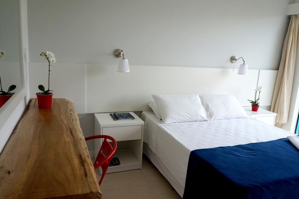 Una habitación de hotel con un escritorio de madera, una cama con ropa de cama blanca, dos lámparas de pared y un ventanal con cortinas.